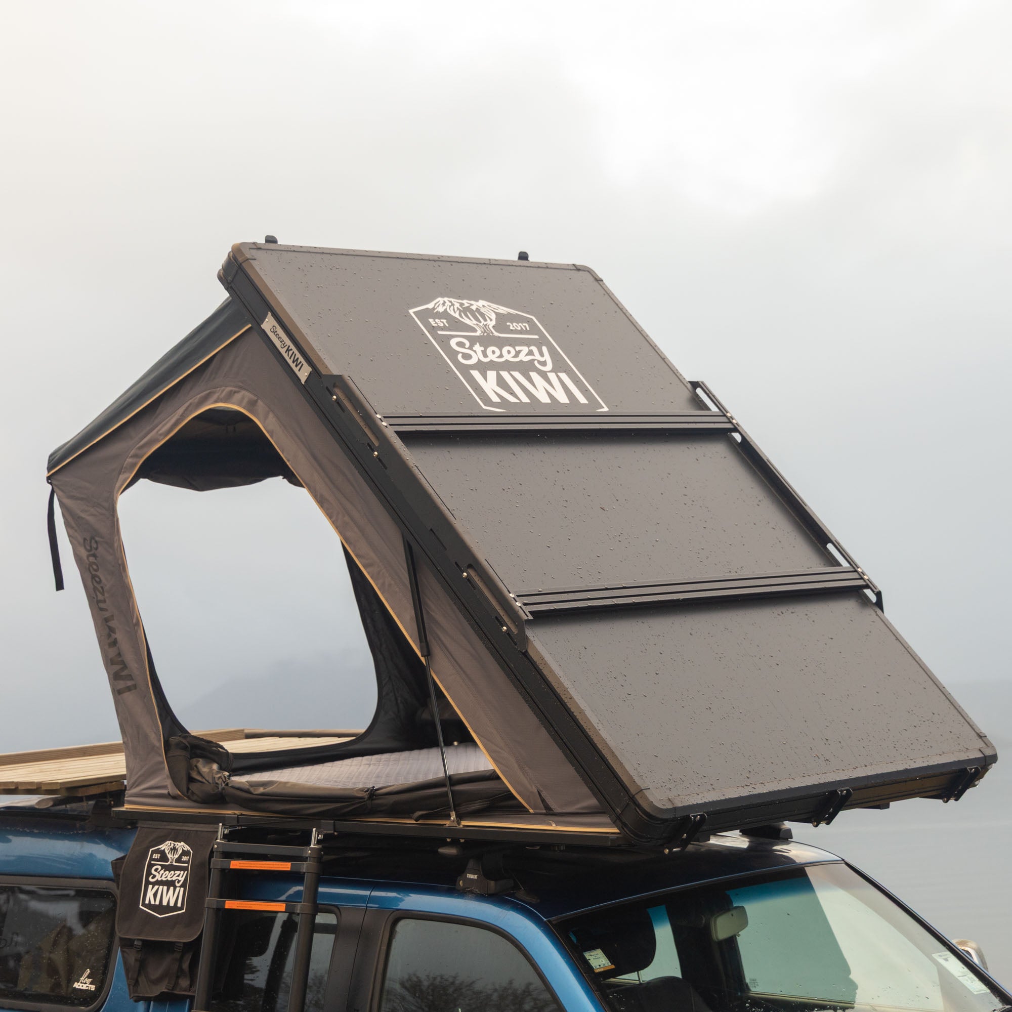 Roof top tent attached to a vehicle with 'Sleazy Kiwi' branding on a cloudy day.